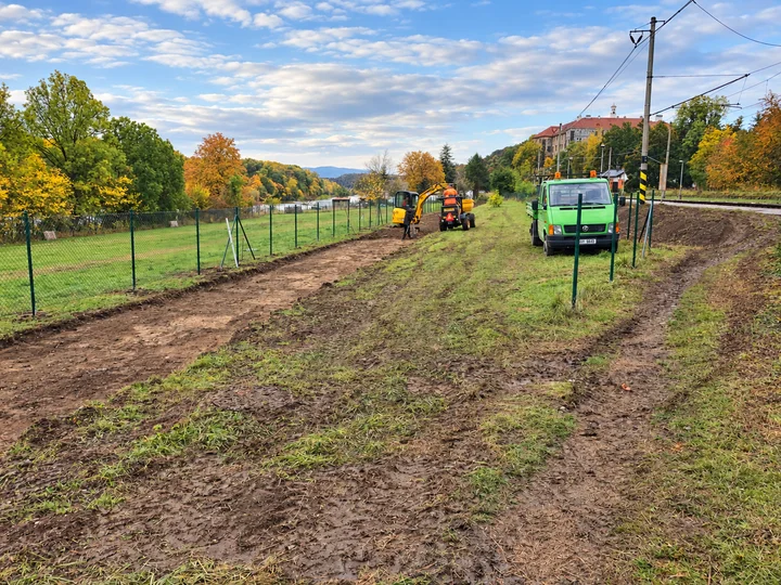 Bagr, dumper a zelený nákladní vůz při úpravě terénu podél oplocení