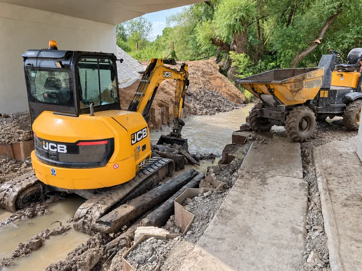 Pásový bagr JCB a dumper při zemních pracích ve vodním toku pod mostem